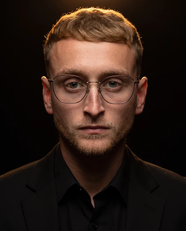Portrait of James Curtis, wearing glasses and a dark blazer, looking directly at the camera against a dark background.