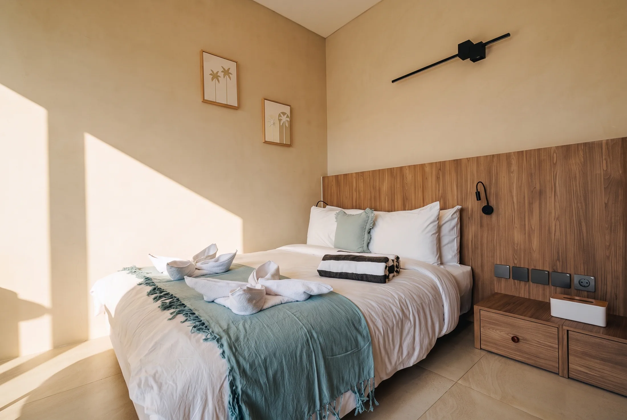 Villa Mares bedroom bathed in warm afternoon light, showing the walnut headboard, folded towels and sage-green throw on the bed